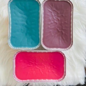 May include: Three rectangular wax melts in foil containers. The wax colours are teal, mauve, and hot pink. The melts are on a white, fluffy surface. The wax has a smooth, slightly textured appearance.