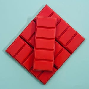 Könnte beinhalten: Leuchtend rote Wachsduftriegel auf hellblauem Hintergrund. Die Wachsduftriegel sind rechteckig mit segmentierten Abschnitten und in einem Rautenmuster angeordnet. Ein Riegel liegt mittig auf der Raute.
