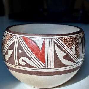 May include: A handcrafted ceramic bowl with a cream-colored base and a decorative band featuring geometric patterns in brown, red, and white. The bowl's rim and base are outlined in brown.