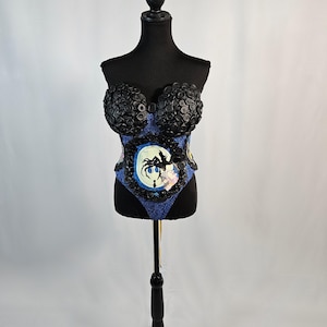 May include: A black corset-style top on a mannequin. The top features black button detailing around the bust and a blue patterned fabric on the bodice. A circular graphic with a silhouette is centered on the front.