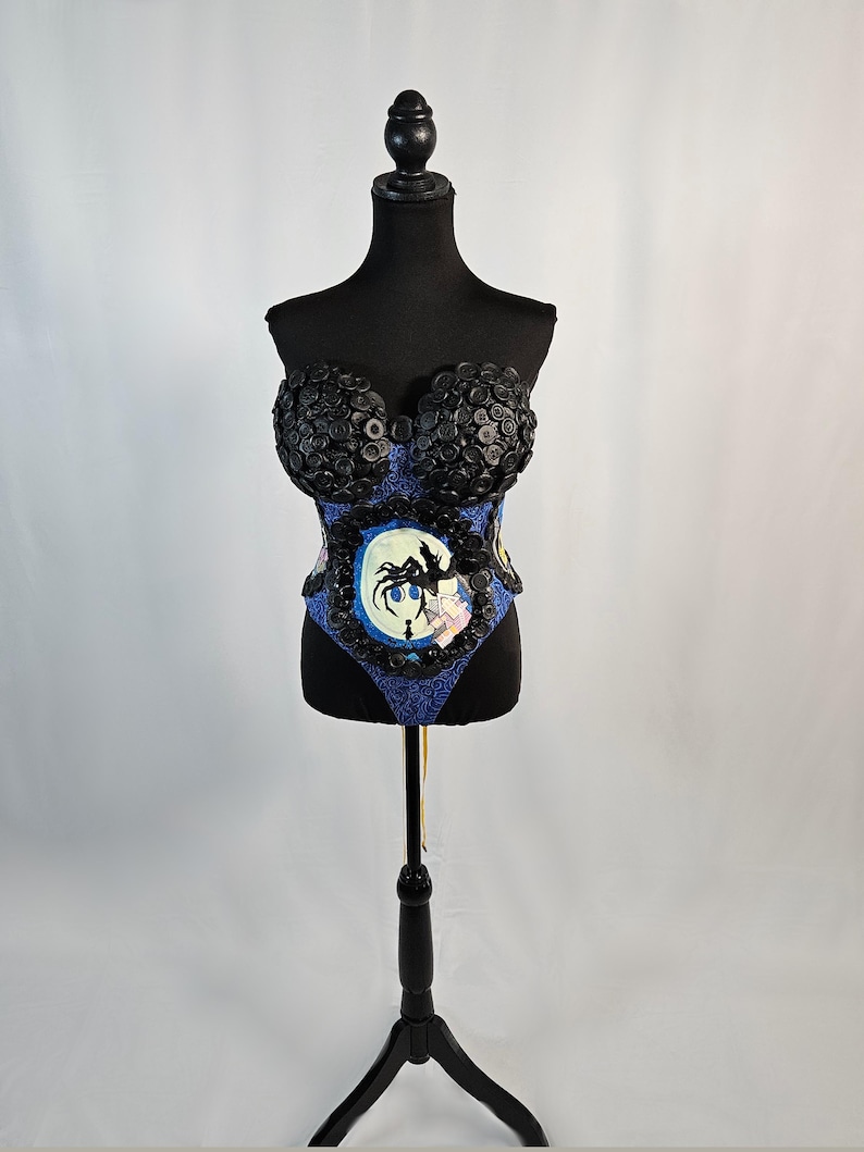 May include: A black corset-style top on a mannequin. The top features black button detailing around the bust and a blue patterned fabric on the bodice. A circular graphic with a silhouette is centered on the front.