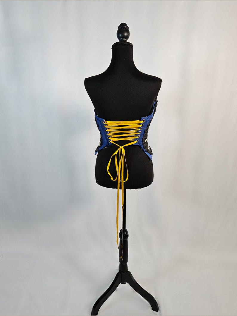 May include: A blue and black corset with yellow lacing is displayed on a black mannequin. The corset has a strapless design and is made of a textured fabric. The mannequin is on a black stand against a white background.