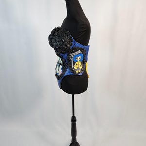 May include: A blue and black corset-style garment with button and character embellishments. The top portion is covered in black buttons, while the midsection features cartoon character designs. The garment is displayed on a black mannequin.