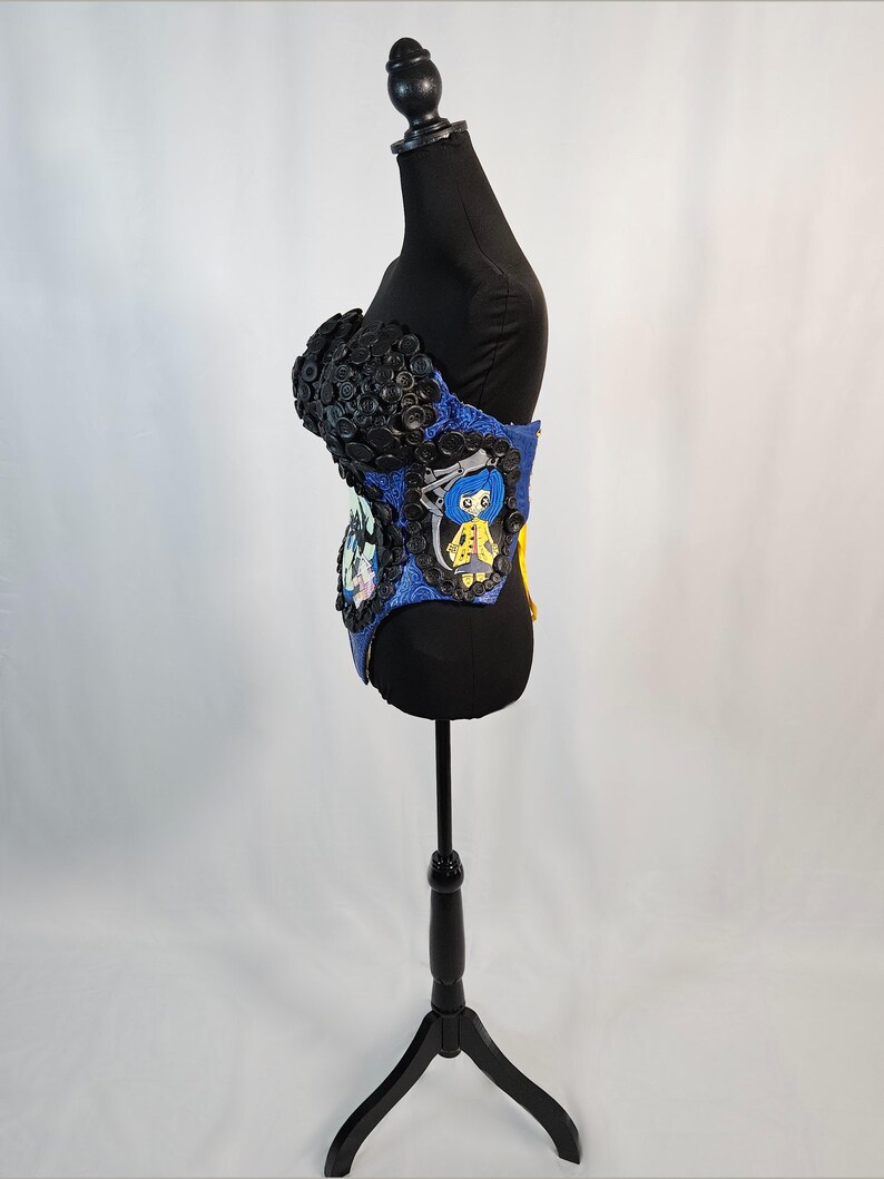 May include: A blue and black corset-style garment with button and character embellishments. The top portion is covered in black buttons, while the midsection features cartoon character designs. The garment is displayed on a black mannequin.
