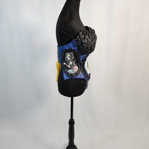 May include: A blue and black corset with a decorative design. The corset features a portrait of a cartoon character and is embellished with black buttons. The corset is displayed on a black mannequin.