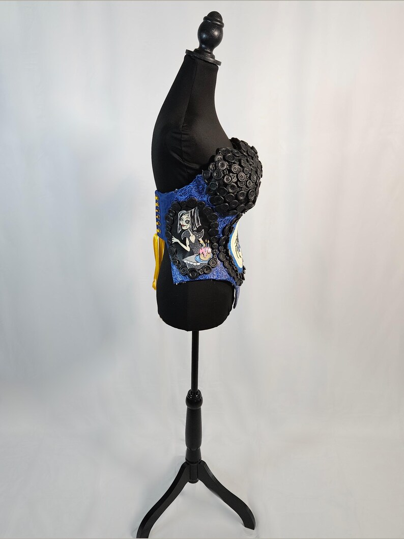 May include: A blue and black corset with a decorative design. The corset features a portrait of a cartoon character and is embellished with black buttons. The corset is displayed on a black mannequin.
