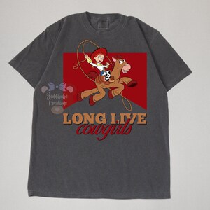 Op de afbeelding: Een donkergrijs T-shirt met een rode, witte en bruine afbeelding van een cartoon-cowgirl die op een paard rijdt. De tekst "Long Live Cowgirls" is in bruin op het shirt gedrukt.