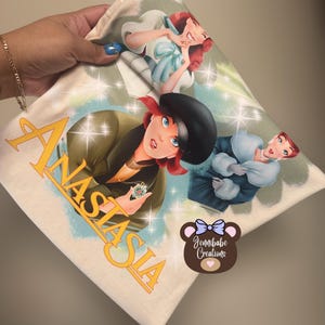 Op de afbeelding: Een wit T-shirt met een kleurrijke afbeelding van drie vrouwen uit de animatiefilm Anastasia. De afbeelding bevat het woord "Anastasia" in een gestileerd lettertype. Het T-shirt heeft ook een klein bruin berenlogo met een roze hart en de tekst "Jennbabe Creations".