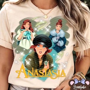 Op de afbeelding: Een beige T-shirt met een grafische print van drie vrouwen uit de animatiefilm Anastasia. De vrouwen dragen kleurrijke jurken en de tekst "Anastasia" is gedrukt in een gestileerd lettertype.