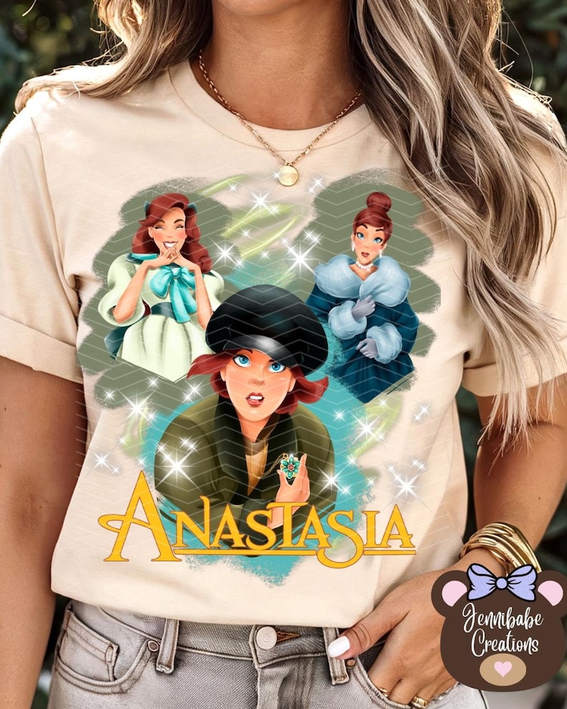 Op de afbeelding: Een beige T-shirt met een grafische print van drie vrouwen uit de animatiefilm Anastasia. De vrouwen dragen kleurrijke jurken en de tekst "Anastasia" is gedrukt in een gestileerd lettertype.