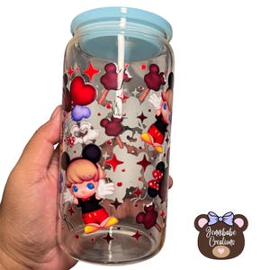 May include: Clear glass tumbler with a light blue lid and straw. The tumbler is decorated with cartoon Mickey Mouse and Minnie Mouse characters, red stars, and ice cream bars. The tumbler is held by a hand.