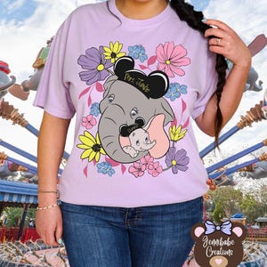 Op de afbeelding: Een lichtpaarse T-shirt met een bloemenontwerp met een grijze olifant en een babyolifant met roze oren. De olifant draagt ​​een zwarte Mickey Mouse-oorkap met de tekst "Mrs. Jumbo" en de babyolifant draagt ​​een zwarte Mickey Mouse-oorkap met de tekst "Dumbo".