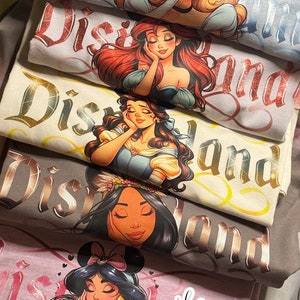 Op de afbeelding: Een stapel t-shirts met Disney-prinsessen. Elk shirt toont een prinsessenafbeelding met het woord "Disneyland" in een decoratief lettertype. De prinsessen zijn onder meer Aurora, Ariel, Belle, Jasmine en anderen. De shirts zijn in verschillende kleuren, waaronder grijs, beige en roze.