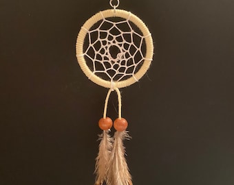 May include: A beige dreamcatcher keychain with a silver keyring. The dreamcatcher features a woven web design within a circular frame. Two wooden beads and brown feathers dangle from the bottom, creating a bohemian aesthetic.