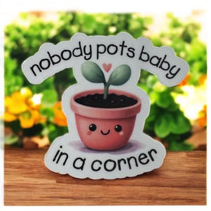 May include: A sticker featuring a cartoon illustration of a potted plant with a smiling face and the text "nobody pots baby in a corner". The plant has two green leaves and a small pink heart above it. The pot is a light reddish-orange color.