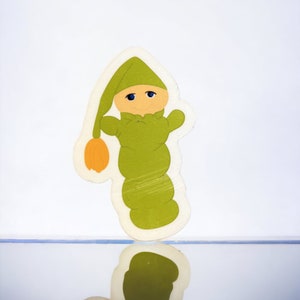 May include: A green felt cutout of a baby wearing a green hat and a yellow blanket. The cutout is reflected in a mirror.