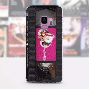 May include: A black and clear phone case with a pink and silver design. The design features a VHS tape with the movie "Clueless" on it. The case has a cutout for the phone's camera.