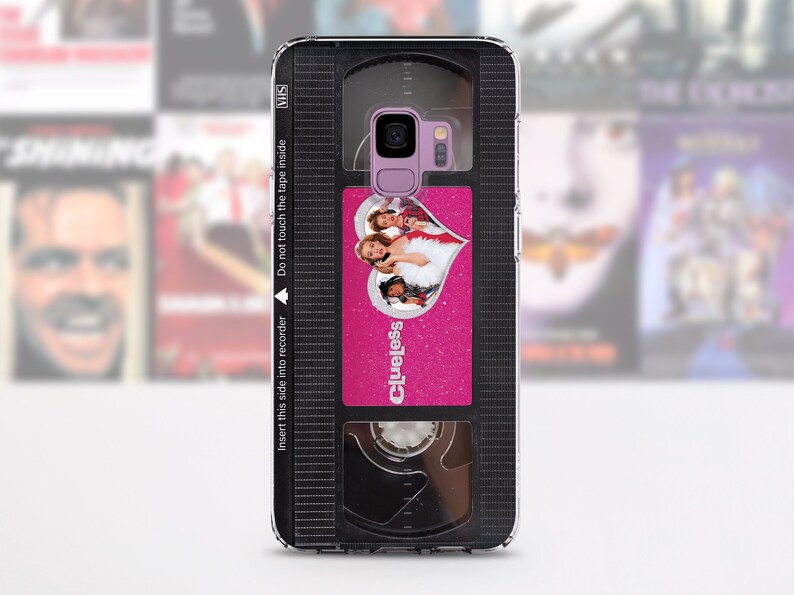 May include: A black and clear phone case with a pink and silver design. The design features a VHS tape with the movie "Clueless" on it. The case has a cutout for the phone's camera.