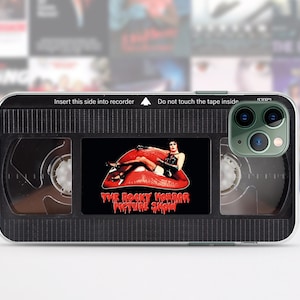 May include: Black phone case with a clear back and a black VHS tape design. The tape has a red lip with a woman in a white outfit sitting on it. The text on the tape reads "The Rocky Horror Picture Show".
