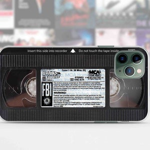 Puede incluir: Una funda de teléfono negra y transparente con un diseño que parece una cinta VHS. La funda tiene una etiqueta en blanco y negro que dice "Regreso al futuro" y "¡Advertencia del FBI!" La funda tiene una parte posterior transparente que muestra las lentes de la cámara del teléfono.