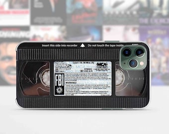 May include: A black and clear phone case with a design that looks like a VHS tape. The case has a black and white label that says "Back to the Future" and "FBI Warning!" The case has a clear back that shows the phone's camera lenses.