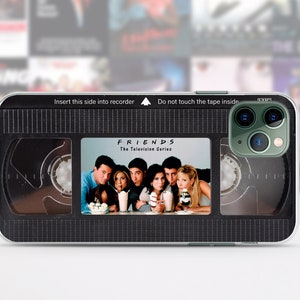 Puede incluir: Una funda de teléfono negra y transparente con un diseño que parece una cinta VHS. La cinta tiene las palabras "FRIENDS The Television Series" y una imagen del elenco del programa. La funda tiene un patrón de cuadrícula en blanco y negro en la parte posterior.