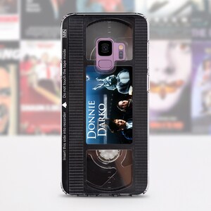 Puede incluir: Una funda para tel&eacute;fono negra y transparente dise&ntilde;ada para parecer una cinta VHS. La funda tiene una ventana transparente que muestra un cartel de pel&iacute;cula para la pel&iacute;cula "Donnie Darko". La funda tiene el texto "Inserte este lado en la grabadora" y "No toque la cinta dentro" impreso en ella.