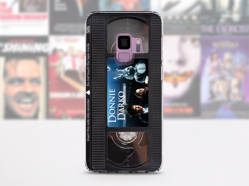 Puede incluir: Una funda para tel&eacute;fono negra y transparente dise&ntilde;ada para parecer una cinta VHS. La funda tiene una ventana transparente que muestra un cartel de pel&iacute;cula para la pel&iacute;cula "Donnie Darko". La funda tiene el texto "Inserte este lado en la grabadora" y "No toque la cinta dentro" impreso en ella.