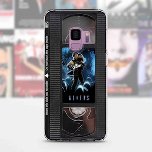 May include: A black and clear phone case with a VHS tape design. The case features a black and white image of the movie "Aliens" on the front. The case has a black and silver camera cutout and the text "Insert this side into recorder" and "Do not touch the tape inside" on the side.