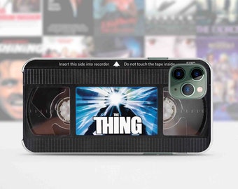 May include: A black and clear phone case with a VHS tape design. The case features the movie title "The Thing" with a blue and white image of the creature from the movie. The case has a black grid pattern and the text "Insert this side into recorder" and "Do not touch the tape inside".