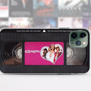 May include: A black and clear phone case with a pink and silver sticker that says "Clueless" and features a heart with a picture of three people. The case is designed to look like a VHS tape.