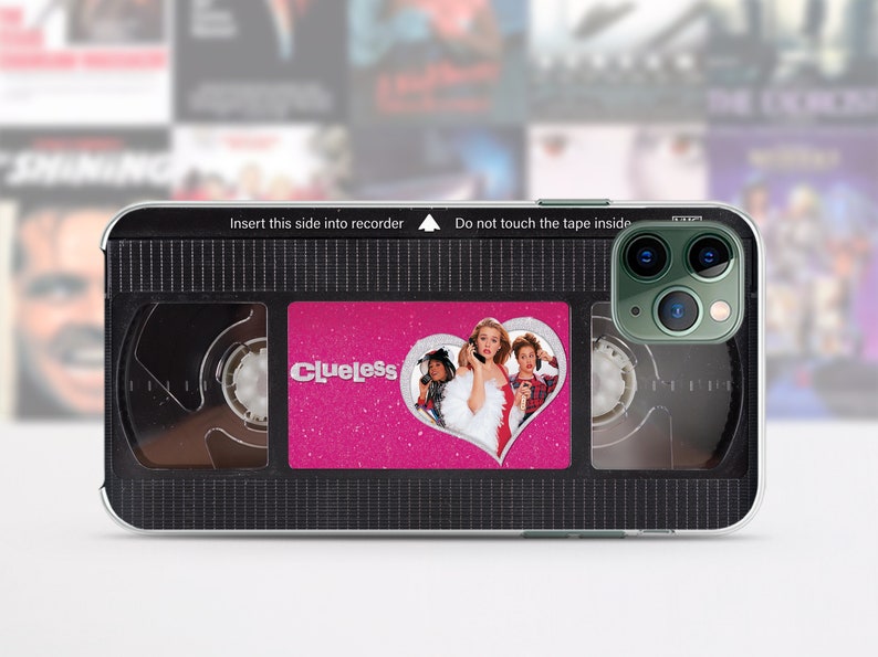 May include: A black and clear phone case with a pink and silver sticker that says "Clueless" and features a heart with a picture of three people. The case is designed to look like a VHS tape.