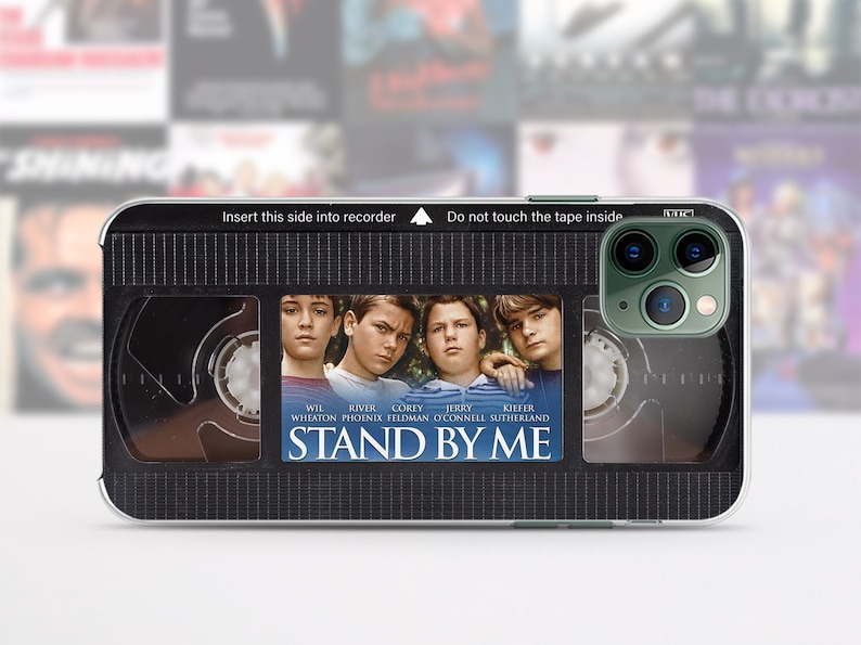 May include: A phone case featuring a VHS tape design with the movie title "Stand By Me." The case has a black background with the movie's poster art and the text "Insert this side into recorder" and "Do not touch the tape inside."