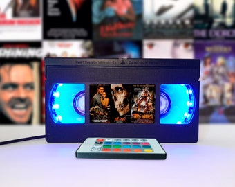 May include: A blue VHS tape shaped lamp with a remote control. The lamp has two clear windows with blue LED lights. The windows display the covers of two horror movies: "Evil Dead" and "Army of Darkness". The text on the VHS tape reads "Insert this side into recorder. Do not touch the tape."
