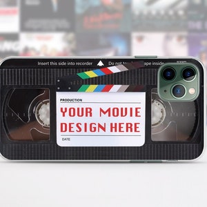 May include: A black and silver phone case with a vintage VHS tape design. The case has a clear window over the camera lens and features the text "YOUR MOVIE DESIGN HERE".