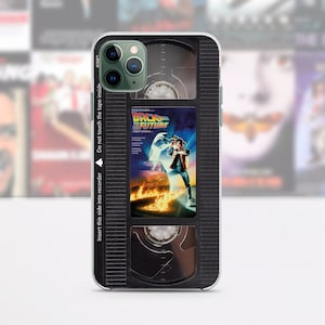 May include: A black phone case with a retro VHS tape design. The case features a clear window showing a movie poster for "Back to the Future". The text on the case reads "Insert this side into recorder" and "Do not touch the tape inside".