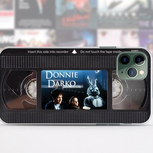 Puede incluir: Carcasa de tel&eacute;fono negra con un dise&ntilde;o que parece una cinta VHS. La cinta tiene las palabras "DONNIE DARKO" y una imagen de un personaje de la pel&iacute;cula. La carcasa tiene un patr&oacute;n de cuadr&iacute;cula en blanco y negro.