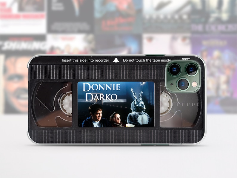 Puede incluir: Carcasa de tel&eacute;fono negra con un dise&ntilde;o que parece una cinta VHS. La cinta tiene las palabras "DONNIE DARKO" y una imagen de un personaje de la pel&iacute;cula. La carcasa tiene un patr&oacute;n de cuadr&iacute;cula en blanco y negro.