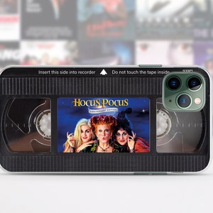 May include: A black and clear phone case with a VHS tape design. The case features the movie "Hocus Pocus" with the three witches on the front. The text "Insert this side into recorder" and "Do not touch the tape inside" is printed on the top of the case.
