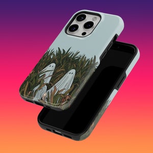 Könnte beinhalten: Eine weiße iPhone-Hülle mit einem Design aus zwei Geistern in einem Feld mit grünem Gras. Die Hülle wird mit einem schwarzen Telefon darin gezeigt.