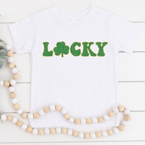 May include: White t-shirt with green and gold "LUCKY" text with a shamrock in the middle.
