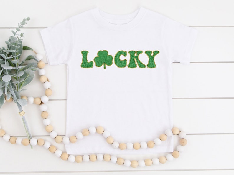 May include: White t-shirt with green and gold "LUCKY" text with a shamrock in the middle.