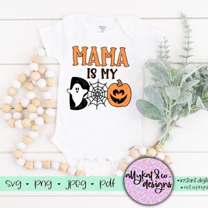 Peut inclure: Une grenouill&egrave;re blanche avec le texte "Mama is my boo" en noir. Un fant&ocirc;me noir et une citrouille orange avec un visage de citrouille sont sur la grenouill&egrave;re. La grenouill&egrave;re est sur une surface blanche avec une cha&icirc;ne de perles en bois et une branche de verdure.