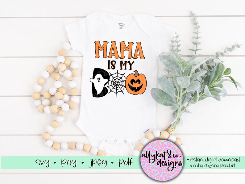 Peut inclure: Une grenouill&egrave;re blanche avec le texte "Mama is my boo" en noir. Un fant&ocirc;me noir et une citrouille orange avec un visage de citrouille sont sur la grenouill&egrave;re. La grenouill&egrave;re est sur une surface blanche avec une cha&icirc;ne de perles en bois et une branche de verdure.