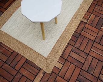 May include: A white, octagonal-shaped table with four wooden legs sits on a natural fiber rug with a brown border. The rug is placed on a dark brown wood floor with a herringbone pattern.