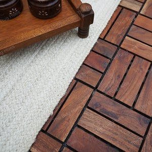 May include: A white woven rug with a wooden tray and two decorative containers on top. The rug is placed on a dark brown wood floor with a herringbone pattern.