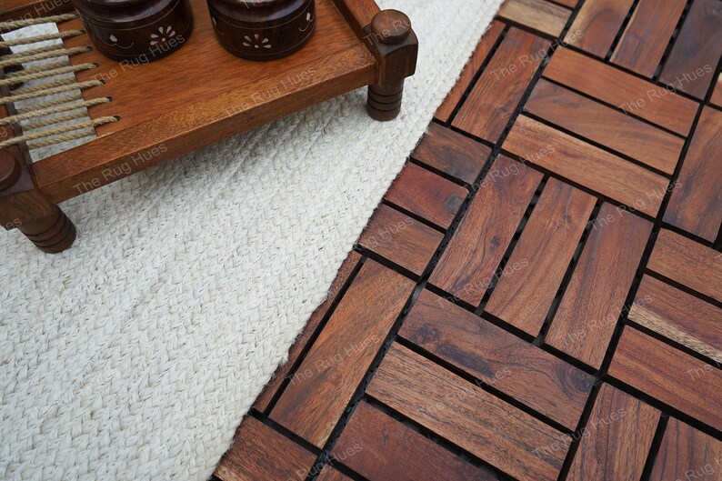 May include: A white woven rug with a wooden tray and two decorative containers on top. The rug is placed on a dark brown wood floor with a herringbone pattern.