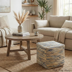 Pode incluir: Cena de sala de estar com uma mesa de centro redonda de madeira, livros e um vaso com capim-dos-pampas. Um pufe cúbico tecido com listras azuis e bege está sobre um tapete de juta. Sofá e cadeiras bege completam o ambiente aconchegante.