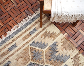 May include: A woven rug with a geometric pattern in beige, brown, and gray tones. The rug has a fringed edge and is partially visible next to a wooden chair and a white blanket with fringe. The floor is made of wooden tiles.