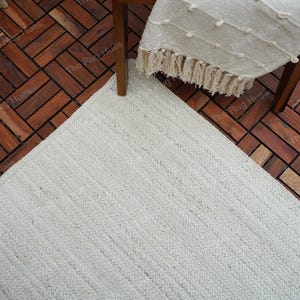 May include: A white woven rug with a textured surface. The rug is placed on a wooden floor with a herringbone pattern.